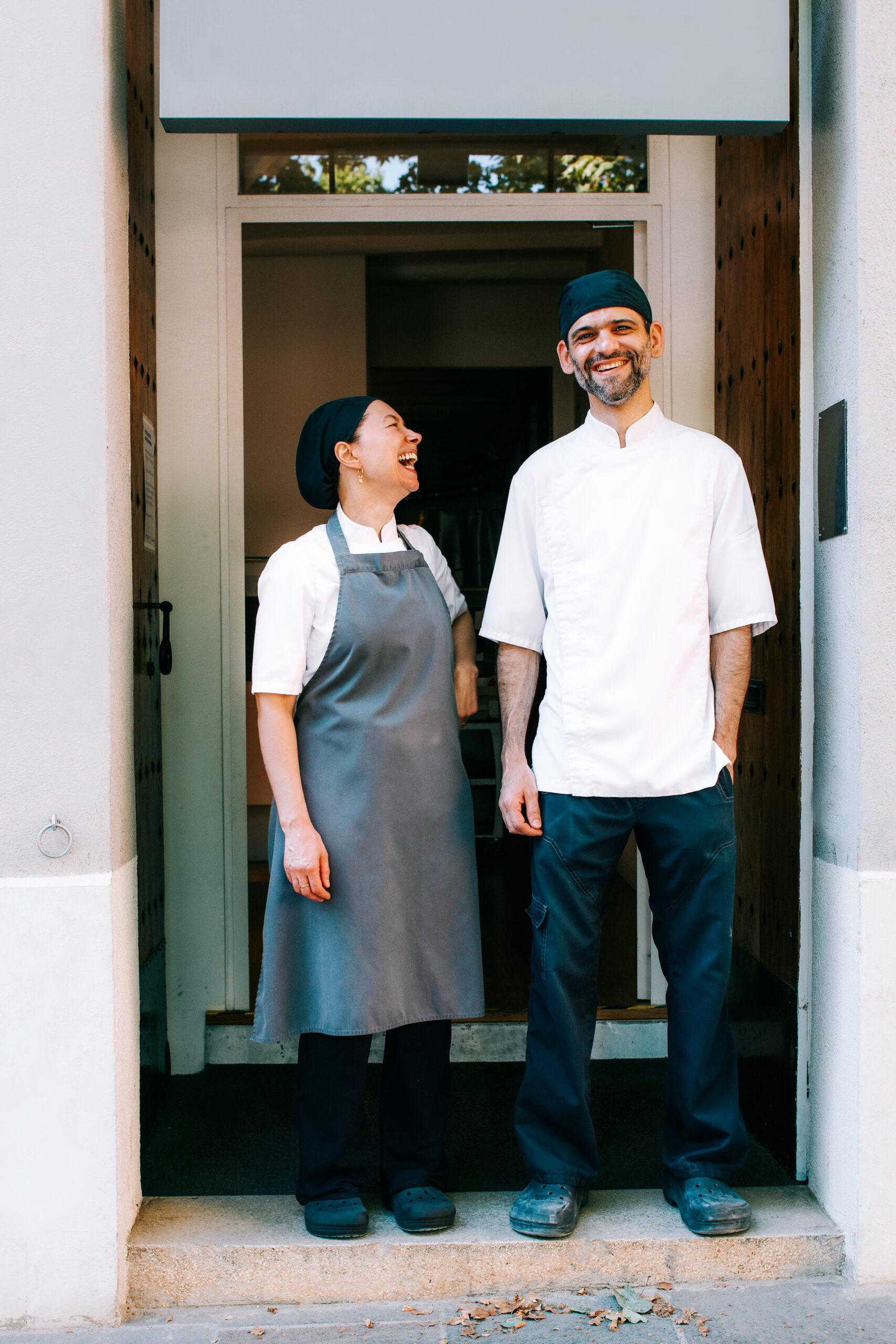 Two restaurant owners, a female wearing an apron smiling at a man with a mustache.