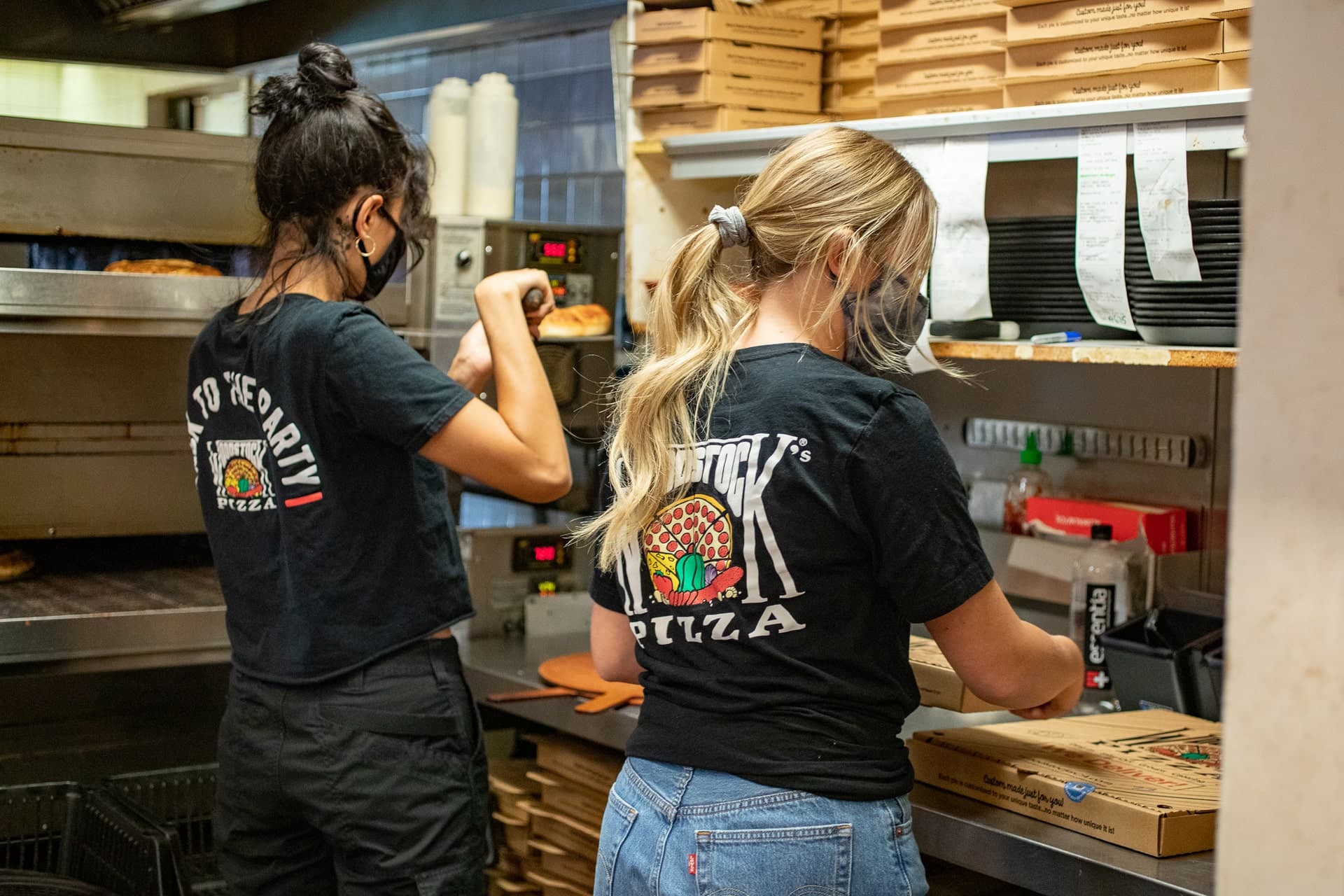 Two female cooks wearing masks, making pizza in the kitchen with order tickets hanging in front of them