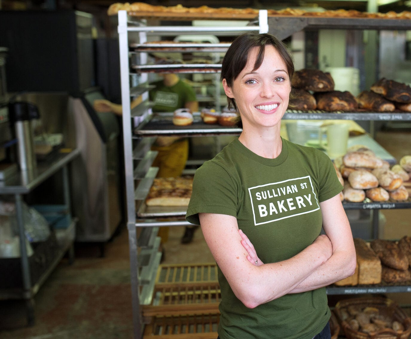 Smiling woman in bakery with bread shelves.