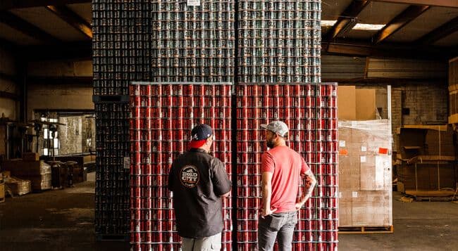 Two men in warehouse with stacked cans.