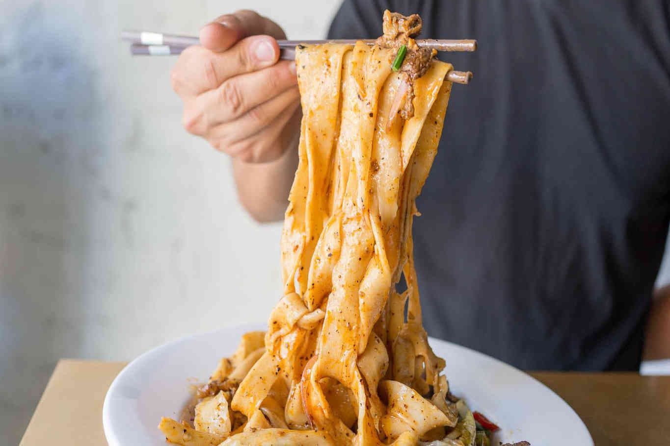 Chopsticks holding wide, spicy noodles above plate.