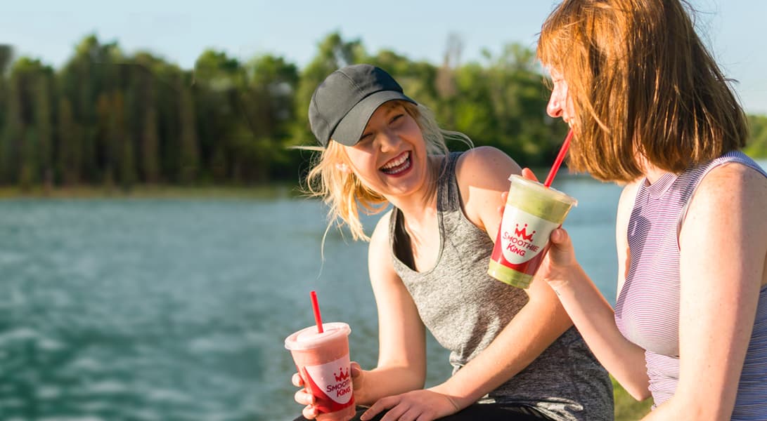 Two friends laughing with smoothies by the lake.