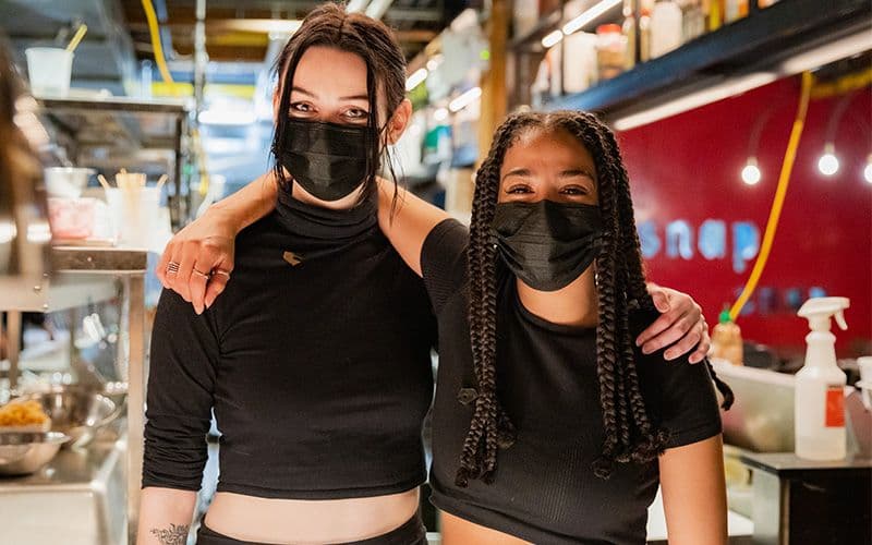Two masked restaurant workers smiling in kitchen.