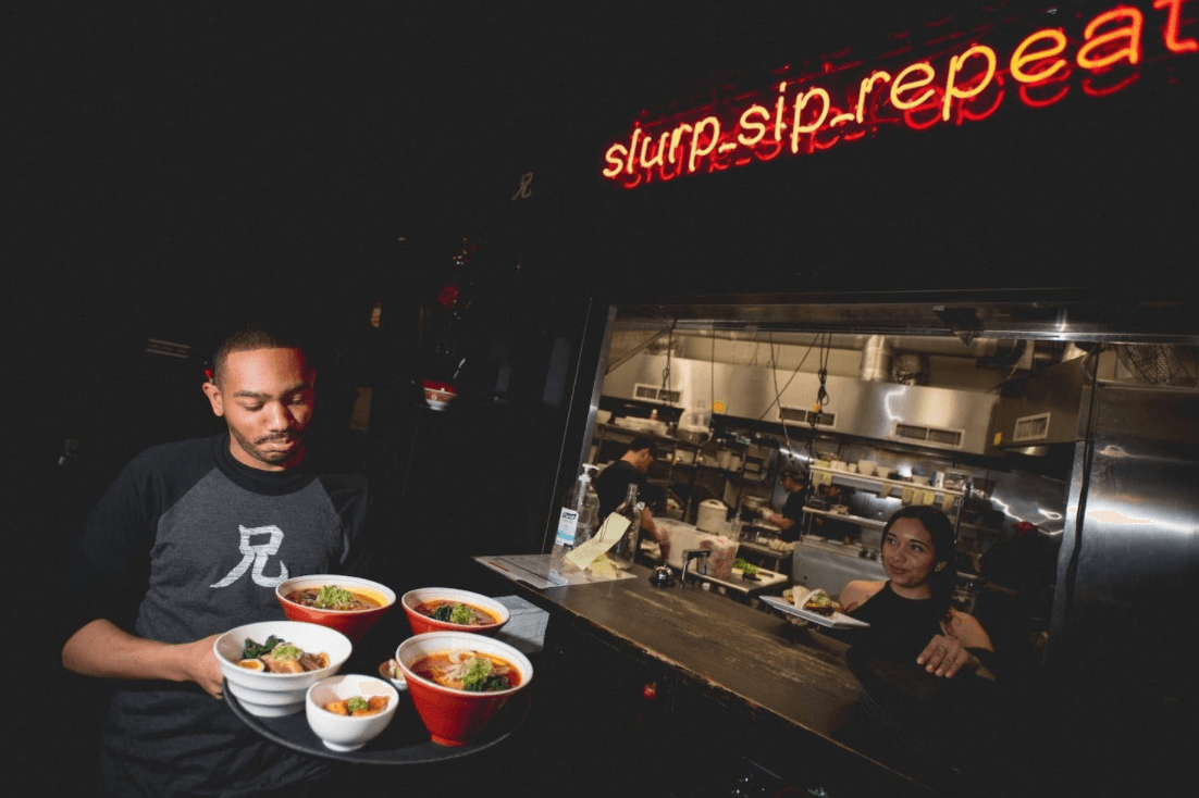 Server with ramen bowls in busy restaurant kitchen.