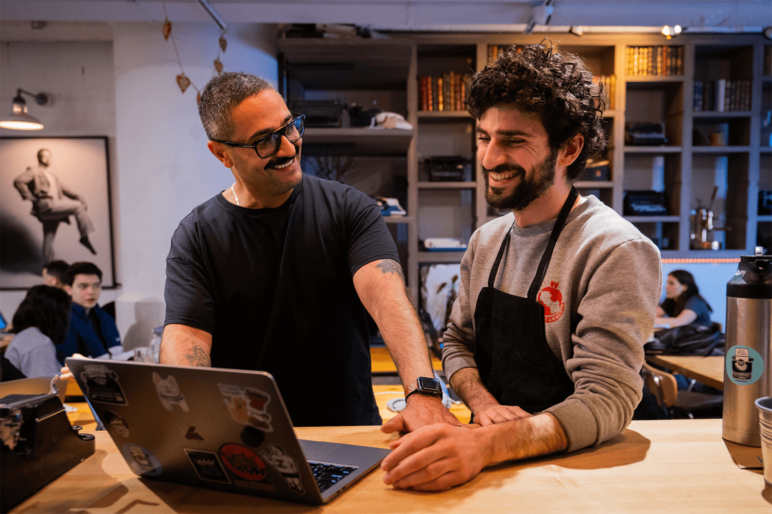 Two men laughing at a laptop in a cafe.