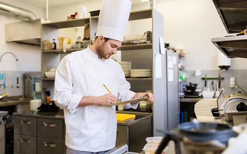 Chef writing in kitchen, wearing white uniform.