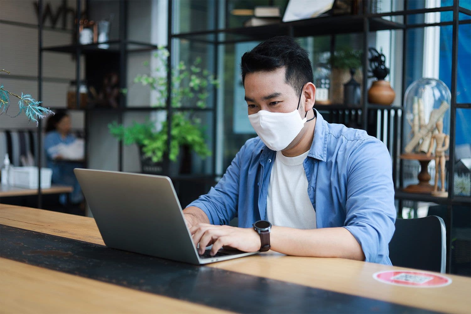 Man with mask working on laptop in cafe.