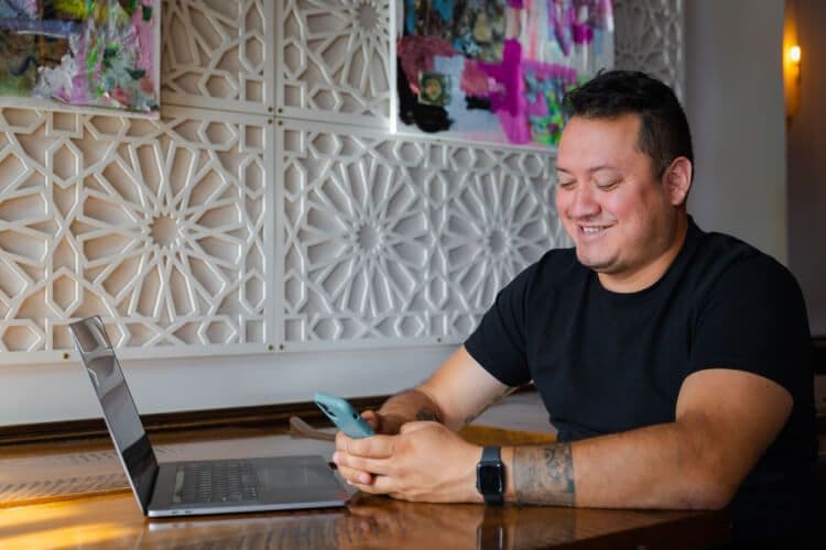Smiling person using phone and laptop at table.