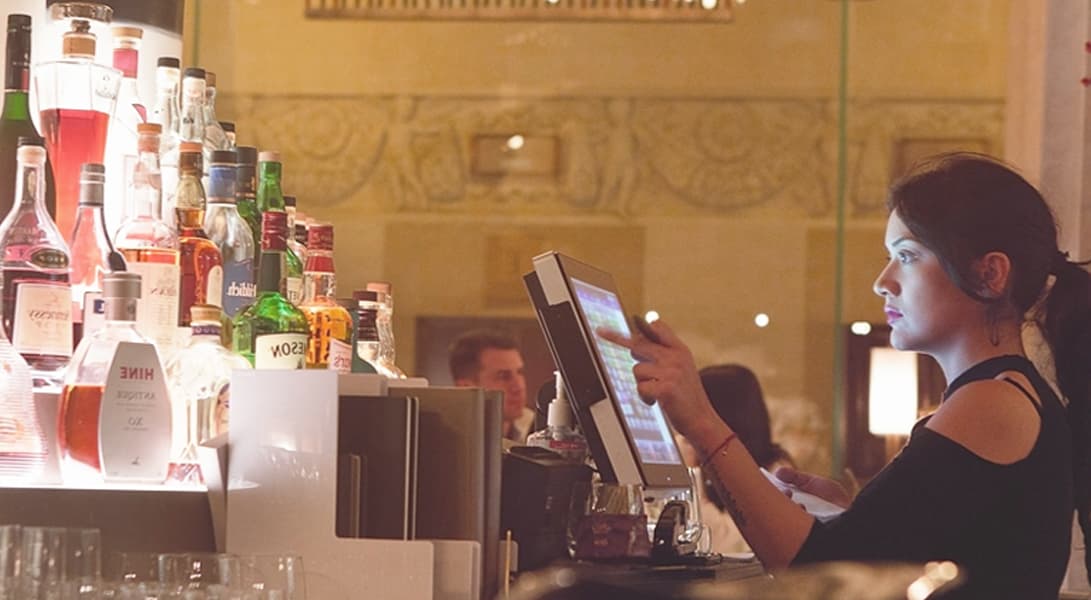Bartender using touchscreen at bar with liquor bottles.
