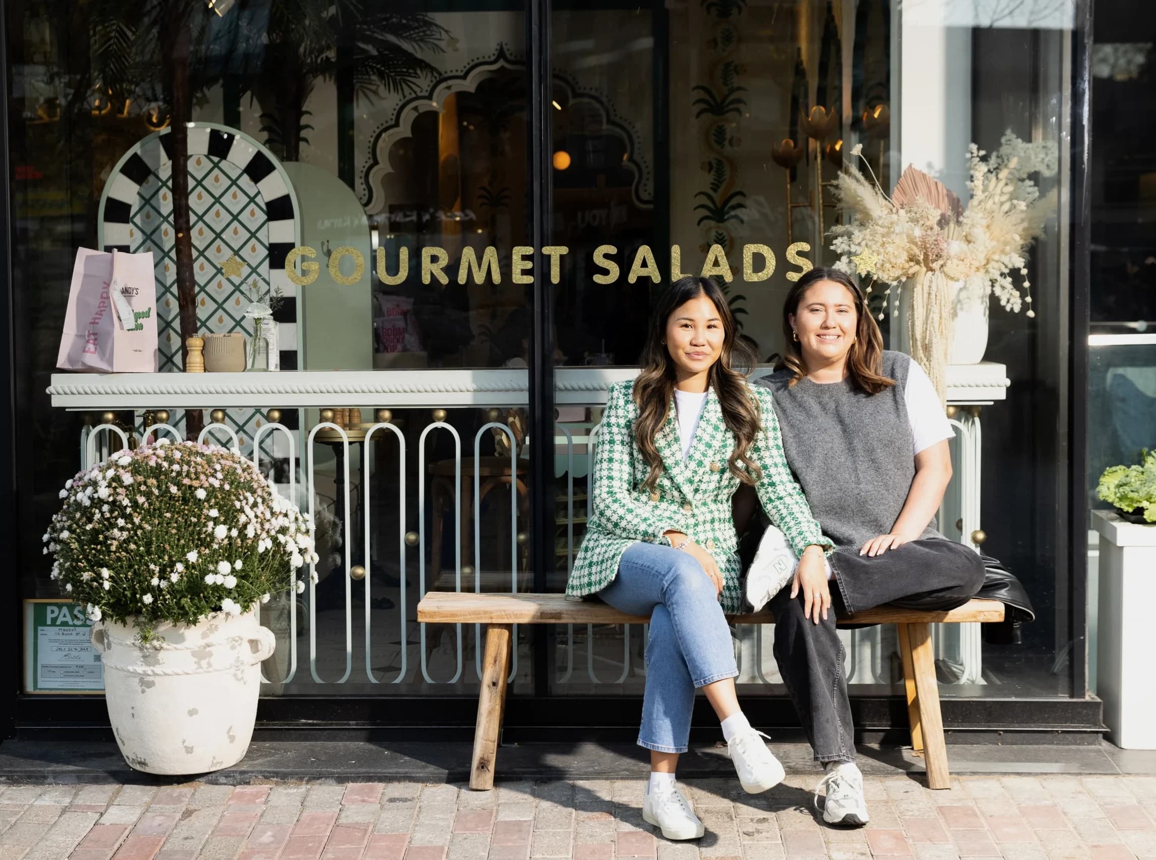 Two smiling women that manage Mandy's Salads sitting on bench outside storefront