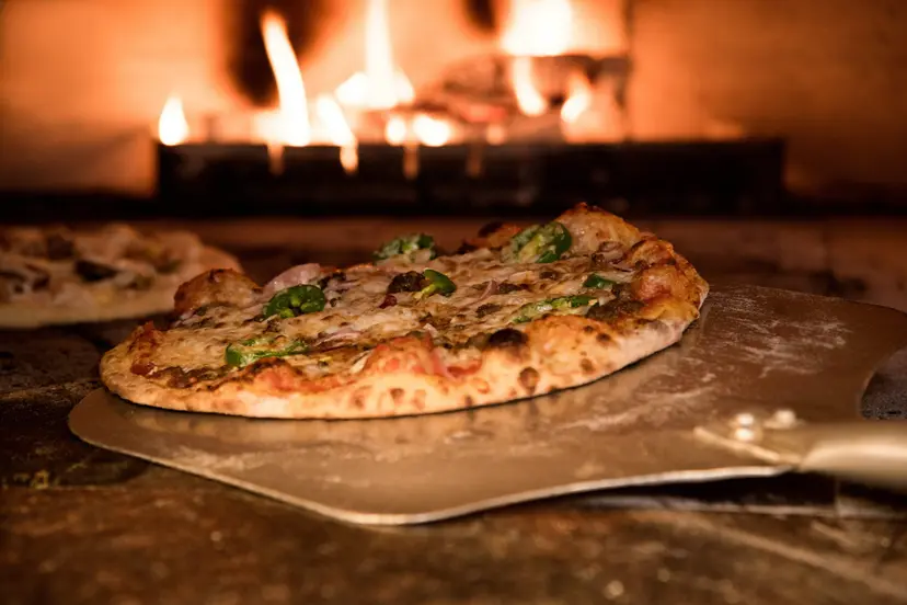 Veggie pizza being placed in a brick oven with flames in the background